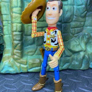 May include: A Woody action figure, wearing a yellow plaid shirt, blue pants, and brown boots. The figure is holding a brown cowboy hat. A sheriff's badge is on the vest. The figure is smiling and has brown hair.