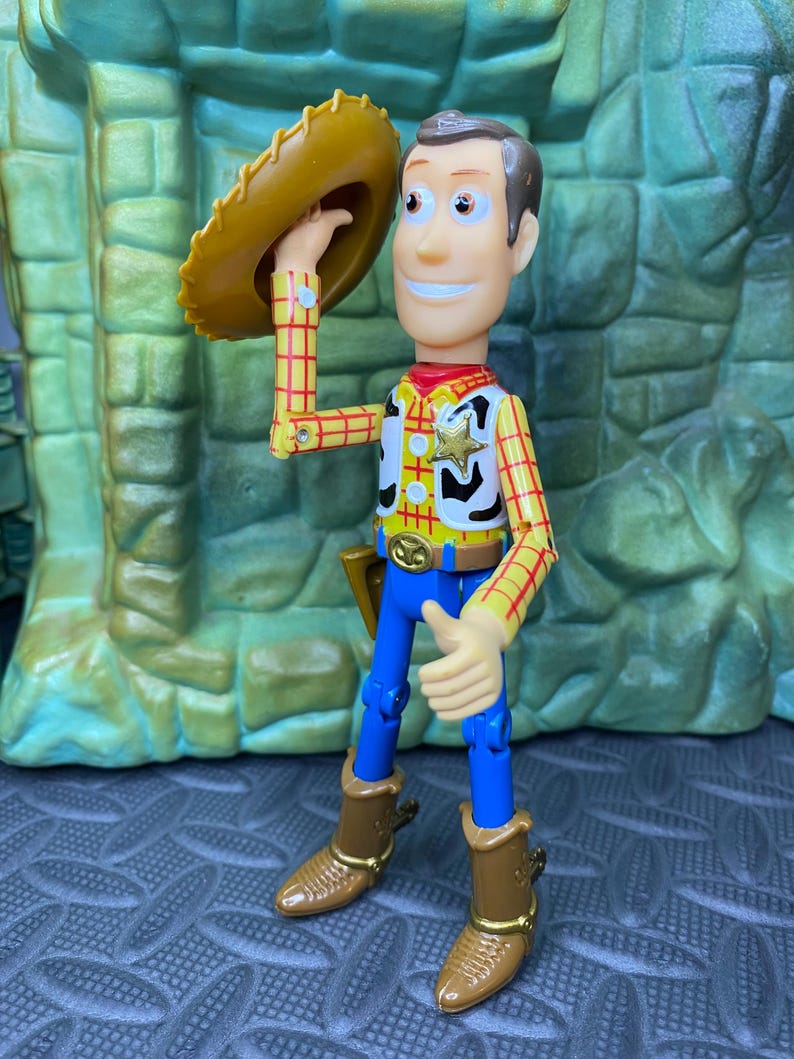 May include: A Woody action figure, wearing a yellow plaid shirt, blue pants, and brown boots. The figure is holding a brown cowboy hat. A sheriff's badge is on the vest. The figure is smiling and has brown hair.