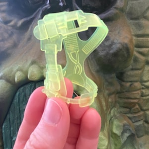 Puede incluir: Un accesorio de juguete de plástico amarillo brillante con un diseño de correa y hebilla. El accesorio está hecho de plástico translúcido y tiene una forma geométrica.