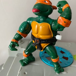Op de afbeelding: Een groene en oranje Teenage Mutant Ninja Turtles-actiefiguur met een zwarte riem en een rode tong die uitsteekt. De figuur is geposeerd met één arm gebogen en de andere arm gestrekt. De figuur staat op een grijs platform.