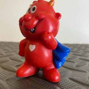 May include: A small red plastic figurine of a cartoon character with a smiling face, two small horns, and a white heart symbol on its chest. The character wears a blue cape attached to its back and stands in an upright position with one arm raised.