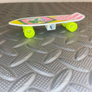 Puede incluir: Un sacapuntas de pl&aacute;stico amarillo y blanco con forma de patineta con un dise&ntilde;o a cuadros rosa y blanco. El sacapuntas tiene un dise&ntilde;o de dibujos animados verde y amarillo en la parte superior.