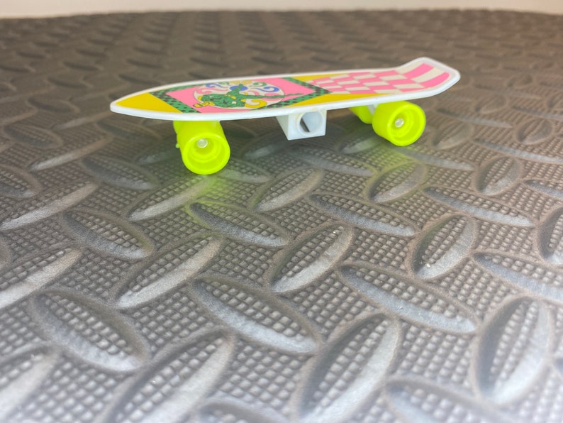Puede incluir: Un sacapuntas de pl&aacute;stico amarillo y blanco con forma de patineta con un dise&ntilde;o a cuadros rosa y blanco. El sacapuntas tiene un dise&ntilde;o de dibujos animados verde y amarillo en la parte superior.