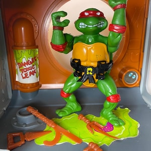 Könnte beinhalten: Eine grüne Teenage Mutant Ninja Turtles Actionfigur mit roter Maske, orangefarbenem Brustpanzer und schwarzem Gürtel mit R-Emblem. Die Figur steht in einem Kanalisations-Spielset mit einem DANGER TOXIC LEAK Schild und orangefarbenen Plastikwaffen.