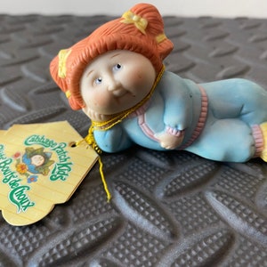 May include: A porcelain Cabbage Patch Kids figurine of a little girl with red hair and a blue and pink outfit. The figurine is lying on its side with its head resting on its hand. The figurine is attached to a yellow tag that reads "Cabbage Patch Kids Little Boots de Choux".