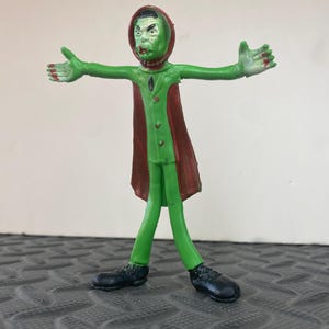 May include: A vintage green plastic monster figure with outstretched arms. The figure has a green face and body, a red cape, and black shoes. The figure is standing on a textured surface.