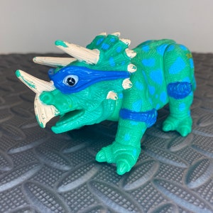 Peut inclure: Un jouet en plastique tricératops vert et bleu avec un masque pour les yeux bleu. Le jouet a des cornes blanches et des accents bleus sur ses jambes.
