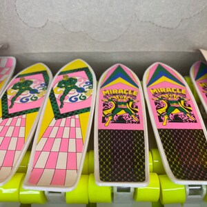Puede incluir: Una caja de seis mini skateboards de dedo con un dise&ntilde;o rosa y amarillo. Cada skateboard tiene una figura verde y las palabras "Miracle Man" impresas en la parte superior.
