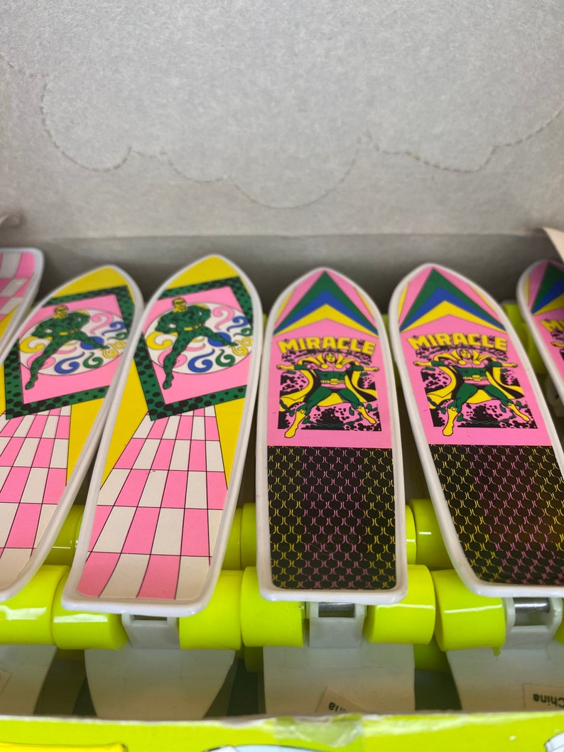 Puede incluir: Una caja de seis mini skateboards de dedo con un dise&ntilde;o rosa y amarillo. Cada skateboard tiene una figura verde y las palabras "Miracle Man" impresas en la parte superior.
