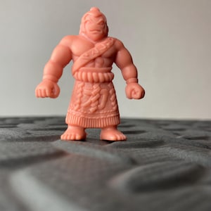 Könnte beinhalten: Kleine, rosa Figur eines muskulösen Kriegers. Die Figur hat einen strengen Ausdruck, einen detaillierten Rock und einen Riemen über der Brust. Das Spielzeug steht auf einer strukturierten Oberfläche.