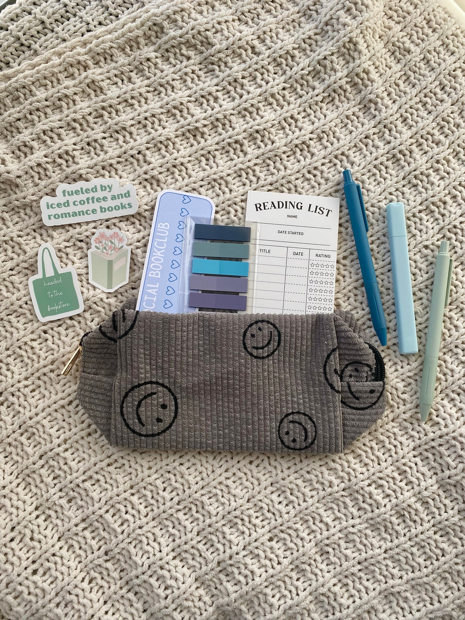 A grey pencil pouch with black smiley faces on it, with a large blue sticker, sticky tabs, and a "reading list" notepad sticking out of it. There are also a couple stickers and pens around it, all sitting on a tan knit blanket.