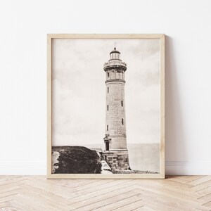 May include: A black and white photograph of a lighthouse on a cliff. The lighthouse is tall and slender, with a white stone exterior and a black roof. The photograph is framed in a light wood frame.