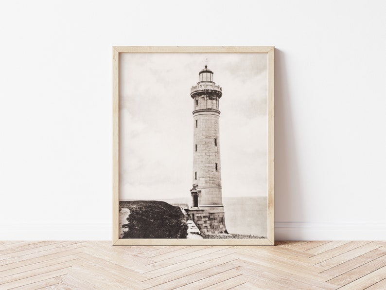 May include: A black and white photograph of a lighthouse on a cliff. The lighthouse is tall and slender, with a white stone exterior and a black roof. The photograph is framed in a light wood frame.
