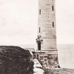 May include: A black and white photograph of a stone lighthouse on a rocky shore. The lighthouse is tall and cylindrical with a small door at the base. The lighthouse is surrounded by a stone wall and a grassy area.