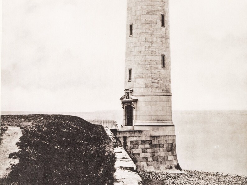 May include: A black and white photograph of a stone lighthouse on a rocky shore. The lighthouse is tall and cylindrical with a small door at the base. The lighthouse is surrounded by a stone wall and a grassy area.