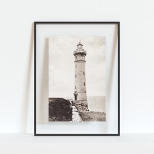 May include: A black and white photograph of a lighthouse on a cliff. The lighthouse is tall and has a white tower with a black roof. The photograph is framed in a black frame.