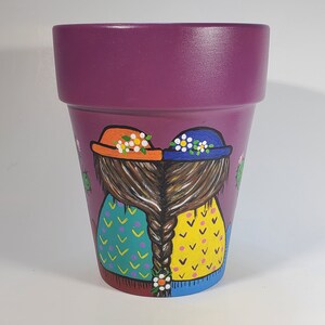 May include: Purple painted terracotta flower pot with two girls with braided hair wearing hats and dresses. The girls are painted in bright colors with yellow, blue, green, orange, and white.