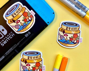 Può includere: Tre adesivi con un'illustrazione di cartone animato di un barattolo di tè al miele e citron. Il barattolo è giallo con un coperchio marrone e ha un'etichetta rossa e bianca con testo coreano che recita "꿀유자차". Il barattolo è circondato da un liquido blu con un mestolo di miele e un cuore rosso. Gli adesivi sono su uno sfondo giallo con due pennarelli gialli e arancioni.