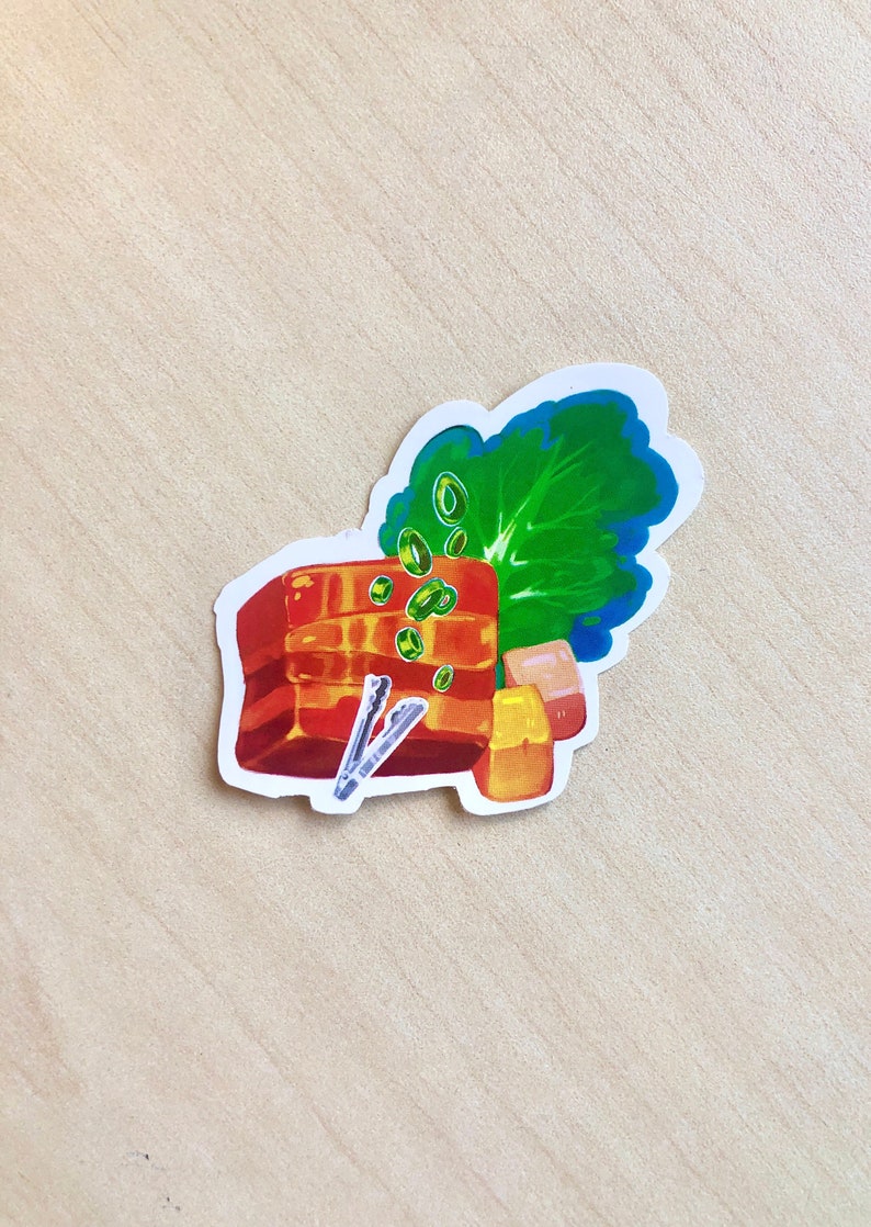 May include: A sticker featuring an illustration of a dish with layers of red-brown meat, green vegetables, and small yellow cubes. The sticker has a white border and is on a light wood surface.