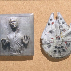 May include: Two Star Wars themed enamel pins. One features Han Solo frozen in carbonite, the other features the Millennium Falcon.
