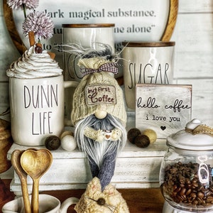 May include: A white ceramic coffee mug with the words "Dunn Life" on it, a white ceramic sugar container with the word "Sugar" on it, a white ceramic creamer with the word "Cream" on it, a white ceramic coffee bean jar with a brown lid, a gnome holding a small coffee mug, and a wooden sign with the words "hello coffee I love you" on it.