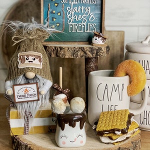 Op de afbeelding: Een houten standaard met twee niveaus met een rustieke houtnerf. De bovenste laag heeft een bord met de tekst "Summer Nights Starry Skies & Fireflies". De onderste laag heeft een witte mok met de tekst "Camp Life" en een s'mores-thema-gnome die een bord vasthoudt met de tekst "S'mores Station".