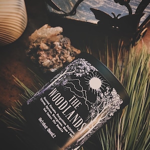 May include: Black candle jar with a white design of trees and a sun. The label reads "THE WOODLANDS" and lists the scent notes: Haunted Forests, Forgotten Folklore, Cypress, Spruce, Evergreen, Moss + Soil, Red Cedar, Fir Needle, October Occult.