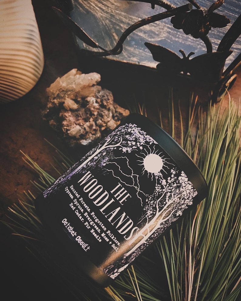 May include: Black candle jar with a white design of trees and a sun. The label reads "THE WOODLANDS" and lists the scent notes: Haunted Forests, Forgotten Folklore, Cypress, Spruce, Evergreen, Moss + Soil, Red Cedar, Fir Needle, October Occult.