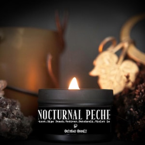May include: A black candle jar with a white label that reads "Nocturnal Peche" and "icot. Ripe Peach. Vetiver. Patchouli. Violet Le" and "October Occult". The candle is lit and the flame is visible.