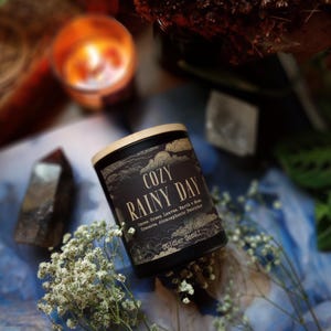 Puede incluir: Una vela negra con tapa de madera, etiquetada como "COZY RAINY DAY". La etiqueta incluye texto sobre aromas y la marca "October Goods". La vela est&aacute; rodeada de flores blancas, cristales y una vela encendida en el fondo. La est&eacute;tica general es acogedora y natural.