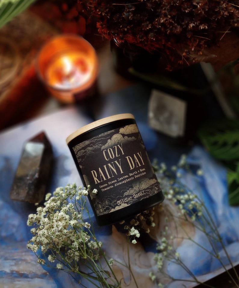 Puede incluir: Una vela negra con tapa de madera, etiquetada como "COZY RAINY DAY". La etiqueta incluye texto sobre aromas y la marca "October Goods". La vela est&aacute; rodeada de flores blancas, cristales y una vela encendida en el fondo. La est&eacute;tica general es acogedora y natural.