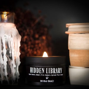 May include: A black candle with the text "HIDDEN LIBRARY" and "October Occult" on the label. The candle is lit and surrounded by books and a jar with a white candle.