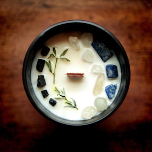 Puede incluir: Un recipiente negro para velas lleno de cera blanca, adornado con peque&ntilde;os cristales, ramitas de vegetaci&oacute;n y una mecha de madera. Los cristales son de tonos claros, azules y amarillos. La vela est&aacute; sobre una superficie de madera marr&oacute;n oscuro.