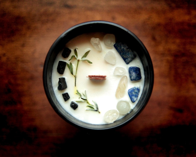 Puede incluir: Un recipiente negro para velas lleno de cera blanca, adornado con peque&ntilde;os cristales, ramitas de vegetaci&oacute;n y una mecha de madera. Los cristales son de tonos claros, azules y amarillos. La vela est&aacute; sobre una superficie de madera marr&oacute;n oscuro.