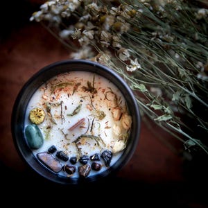 May include: A black candle jar with a white wax candle inside. The candle is decorated with dried flowers, herbs, and small stones.