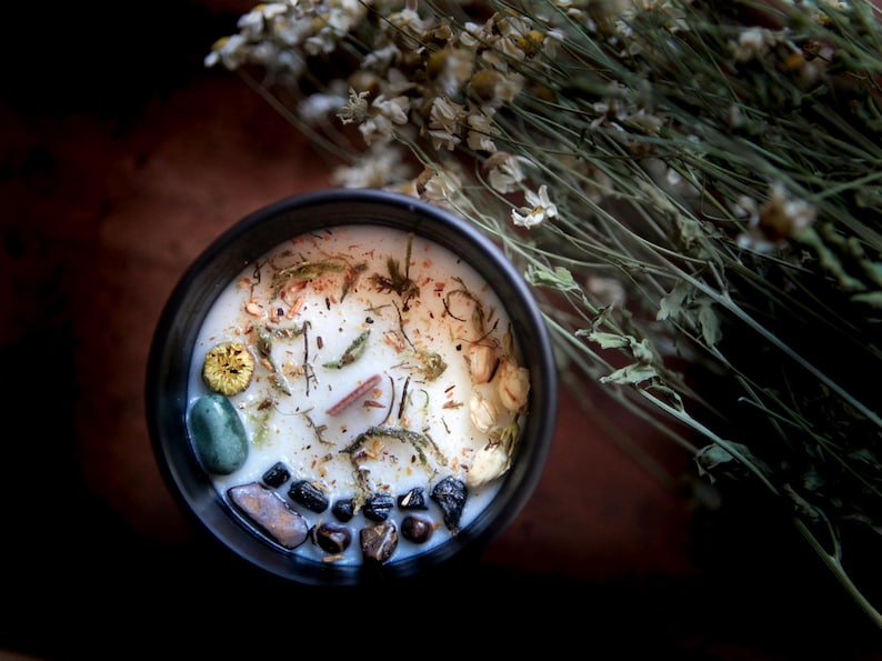 May include: A black candle jar with a white wax candle inside. The candle is decorated with dried flowers, herbs, and small stones.