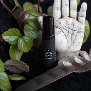 May include: A black roll-on bottle with a white label that says "HEXE" on it. The bottle is sitting on a black surface with a feather and a white plaster hand with lines drawn on it.