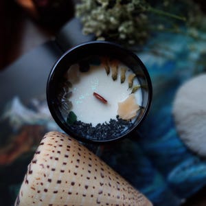 May include: A black candle jar filled with white wax, decorated with small stones, dried flowers, and a wooden wick. A beige seashell with brown spots is in the foreground. The candle is on a blue and white patterned surface.