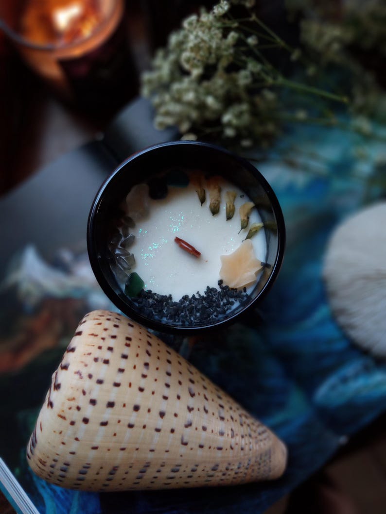 May include: A black candle jar filled with white wax, decorated with small stones, dried flowers, and a wooden wick. A beige seashell with brown spots is in the foreground. The candle is on a blue and white patterned surface.
