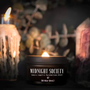 May include: A lit candle in a dark brown tin with the text "MIDNIGHT SOCIETY" and "October Occult". The candle's scent is described as Embers, Campfire Marshmallows, and Ghost. Other candles and crystals are in the background.