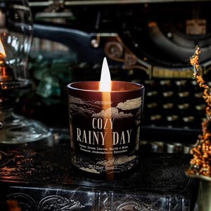 Puede incluir: Una vela encendida, de color marr&oacute;n oscuro, en un tarro de cristal con el texto "COZY RAINY DAY". La vela tiene una llama blanca y un dise&ntilde;o decorativo de nubes. La vela est&aacute; sobre una superficie oscura con una m&aacute;quina de escribir vintage al fondo.