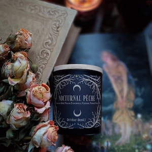 May include: A black candle with a wooden lid, labeled "Nocturnal Pêche" with scent notes. The candle sits near dried peach-colored roses and an open book. A lit candle and a book are in the background.