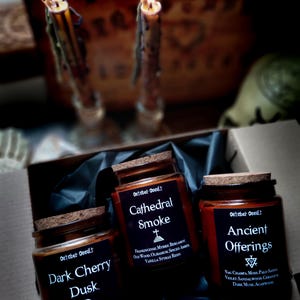 May include: Three glass jar candles with cork lids, labeled "Dark Cherry Dusk", "Cathedral Smoke", and "Ancient Offerings". The labels list scent descriptions. Lit candles are in the background, creating a mystical ambiance.