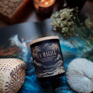 May include: A black candle with a wooden lid, labeled "SEA MAGICK" with ocean-themed imagery. The candle is surrounded by a seashell, a white coral-like object, and dried flowers. The background features a blue and white ocean-themed design.