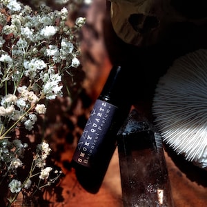 May include: A black roll-on bottle labeled "Smoky Quartz Botanical Perfume" next to a smoky quartz crystal. White flowers and a skull are in the background. The items are arranged on a wooden surface.