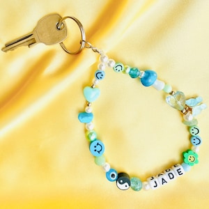 May include: A beaded keychain with a key attached. The keychain features a variety of beads in shades of blue, green, and white, including smiley faces, hearts, and the name "JADE". The background is a yellow satin fabric.