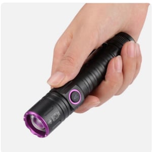May include: A black flashlight with a purple bezel and button is held in a hand. The flashlight has a ribbed body and a circular button with a purple outline. The lens is also purple.