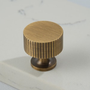 May include: A round, antique brass cabinet knob with a grooved cylindrical body and a circular base. The knob has a brushed finish and is set against a white marble surface. This hardware is suitable for drawers and cabinets.