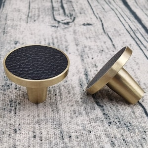 Peut inclure: Deux boutons de cabinet ronds en cuir noir avec une base en laiton. Les boutons ont une surface en cuir texturée et une base en laiton lisse.