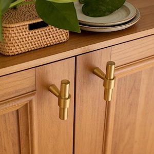 May include: Close-up of a wooden cabinet with two brass-colored T-shaped handles. The cabinet doors are made of light-colored wood. A woven basket and plates are visible on top of the cabinet.